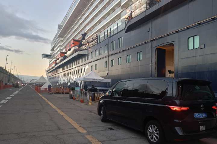 Large cruise ship docked with tents and a black van nearby under a cloudy sky.
