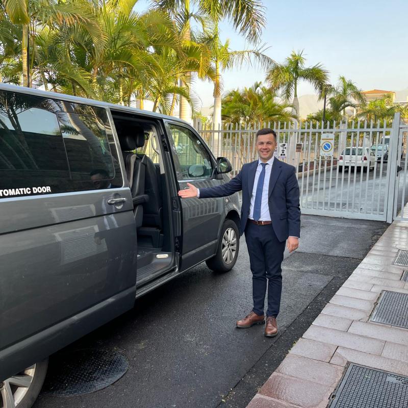 Man in suit standing by a van with an open sliding door, palm trees in the background.